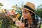 Portrait of pretty woman in a straw hat sitting in the garden, sniffing the scent of roses from a bush. Summer gardening