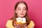 Portrait of pretty young girl, looks at plate with piece of birthday cake  over pink background, wants to eat tasty