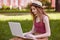 Portrait of pretty young woman sitting on green grass in park with laptop on legs, spending summer day working outdoor, using