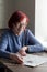 Portrait of red haired elderly woman in glasses, sitting at table in her house, solving crossword puzzle. Cognitive rehabilitation