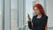 Portrait of a red-haired woman holding a mobile phone in her hand looking gloomily out the window. Angry woman in the