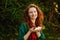 Portrait of a red-haired woman in the park. A curly-haired woman near the green fir branches holds small rose petals in her hands