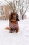 Portrait of Red longhaired dachshund sitting on snow in winter park, little fluffy doggy wearing winter clothin