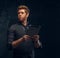 Portrait of a redhead man in formal wear holding a tablet in studio against a dark textured wall