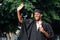 Portrait of rejoicing black guy graduate from university stand outdoors near trees waving hand and holding higher