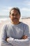 Portrait of retired biracial senior man with arms crossed at beach against sky during sunny day