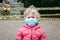 Portrait of sad Caucasian girl in face mask on closed playground outdoor. Kids play area locked with yellow caution tape in