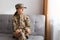 Portrait of sad depressed pensive woman soldier returning home from army, wearing camouflage uniform and cap, sitting on gray