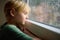 Portrait of a sad preteen boy standing at the window. Child watching the rain outside. Post-traumatic disorder. Accommodation of