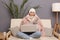 Portrait of sad upset frozen woman wearing cap, gloves, scarf and coat sitting in living room, holding smart phone in hands, holds