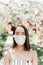 Portrait of sad young woman in protective medical face mask with flowers near blooming tree in spring time