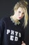 A portrait of a sassy blond woman with blue eyes and Phys Ed black shirt and necklace.