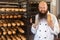 Portrait of satisfied adult baker with long beard in white uniform standing in bakery and holding phone and fresh bread, looking