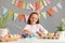 Portrait of satisfied delighted little girl wearing rabbit ears sitting at table painting eggs preparing for Easter, waiting for