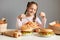 Portrait of satisfied little girl with pigtails sitting at table with big variety of junk food, eating pasta with satisfied