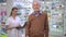 Portrait of satisfied senior Caucasian man posing with shopping bag in pharmacy looking back at blurred woman with