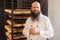 Portrait of satisfied young adult baker with long beard in white uniform standing in his workplace, near shelves with bread at the
