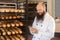 Portrait of satisfied young adult businessman baker with long beard in white uniform standing in bakery and have online order by