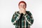 Portrait of scared bearded guy wearing hat and plaid shirt covering mouth, while standing isolated over white background