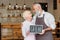 portrait of senior coffee shop owners in aprons holding open chalkboard together