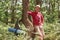 Portrait of senior man standing near tree over forest background. Mature man with rucksack and rug stops to have rest after long