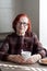 Portrait senior woman with red hair, wearing eyeglasses, in kitchen, sitting at table with cup of tea. elderly woman looks into