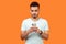 Portrait of serious attentive brunette man with beard in white t-shirt using cellphone. indoor studio shot isolated on orange