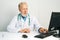 Portrait of serious mature adult male doctor wearing white coat with stethoscope working on desktop computer sitting at