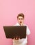 Portrait of shocked guy in white hoodie stands on pink background with laptop in hand, looks in screen with astonishment.Surprised
