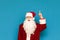 Portrait of shocked Santa Claus on blue background, showing thumbs up at copyspace, looking into camera with surprised face.
