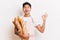 Portrait shot of Asian man baker holds bunch of french baguettes in paper bag makes ok gesture looks satisfied wears