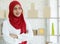 Portrait shot of cute smiling young teenage Muslim woman wearing red hijab sitting on a chair and scanning boxes with barcode