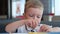 Portrait small blond caucasian boy in white t-shirt sits at table in fast food restaurant and eats junk food. Child dips