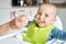 Portrait Of Smiling 8 month Old Baby Boy At Home In High Chair Being Fed Solid Food By Mother With Spoon