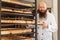 Portrait of smiling adult baker with long beard in white uniform standing in his workplace and pushing baking tray of bread in the
