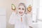 Portrait of smiling attractive elderly woman holding two halves of avocado, with nourishing facial clay mask, posing