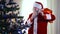 Portrait of smiling bearded Santa admiring decorated Christmas tree in living room standing with red gift bag. Happy old