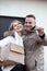 Portrait of smiling couple in casual upper garments stand embracing, holding keys and paperboard box in front of house.