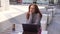 Portrait of smiling dreamy creative pensive young woman sitting with laptop at table in outdoor cafe terrace in sunny