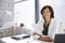 Portrait Of Smiling Female Doctor Wearing White Coat With Stethoscope Sitting Behind Desk In Office