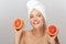Portrait of smiling girl without makeup with white towel on head holding halves of grapefruits near face happily looking