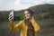 Portrait of smiling girl taking selfie on smartphone on mountain landscape background. Young woman in yellow park jacket enjoys