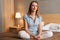 Portrait of smiling peaceful woman relaxing on lotus position sitting on bed in cozy bedroom and looking at camera.