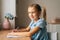 Portrait of smiling pretty primary girl child writing doing homework sitting at home table by window looking at camera.