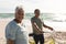 Portrait of smiling retired senior multiracial couple wheeling bicycle at beach on sunny day