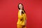 Portrait of smiling stunning young woman in yellow fur sweater pointing thumb aside isolated on bright red wall