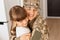 Portrait of smiling young adult soldier woman wearing camouflage uniform and cap posing with her daughter, hugging kid with