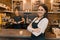 Portrait of smiling young female coffee shop owner, confident woman with arms crossed standing at the counter with barista working