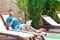 Portrait of smug handsome bearded young adult man in blue t-shirt and shorts lying on cozy sunbed with laptop on poolside and