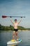 Portrait of a sporty young man standing on the water on a sup boardt raises his hands with an oar and poses for the camera.